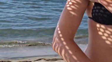 Cilde güneş kremi sürmeye yakın çekim. SPF koruması. Plaj şemsiyesinin gölgesindeki kadın vücuduna güneş kremi sürüyor. Parlak gölgeler. Güvenli bronzlaşma. Plaj tatili