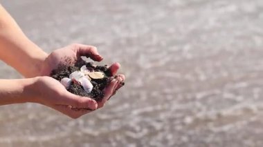 Bir avuç kabuk ve deniz kumu tutan kadın ellerinin yakın çekimi. Denizde tatil, deniz kenarında tatil. Elinde deniz kabukları tutan bir kız. Eller deniz dalgalarının arka planında. Plaj