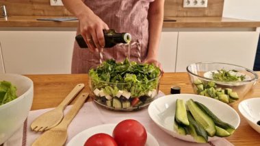 Önlüklü kız bir şişe zeytinyağı açar ve cam salata kasesinde salatanın üzerine döker. Yağ ekledikten sonra, kız salatayı ahşap spatula ile karıştırıyor. Elleri yakın çekim. Kadın mutfakta salata hazırlıyor.