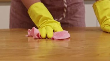 Temizlik şirketinden bir kadın odayı temizliyor. Önlüklü ve eldivenli tanınmayan bir kadın masanın tozunu alıyor. Sarı lastik eldivenli ellerinin yakın çekimi.