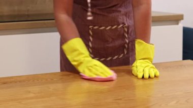 Temizlik şirketinden bir kadın odayı temizliyor. Önlüklü ve eldivenli tanınmayan bir kadın masanın tozunu alıyor. Sarı lastik eldivenli ellerinin yakın çekimi.