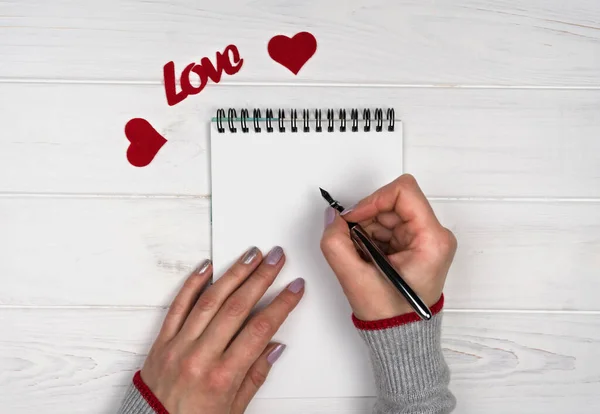 Woman\'s hand writes love message on notepad. Flatlay composition. Love romantic message for Valentine\'s Day. Valentine\'s Day holiday concept. Top view.