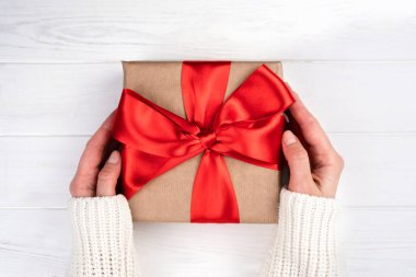 Woman's hands holding gift or present box with red bow on white wooden background. Close-up. Top view. Flatlay. Selective focus.