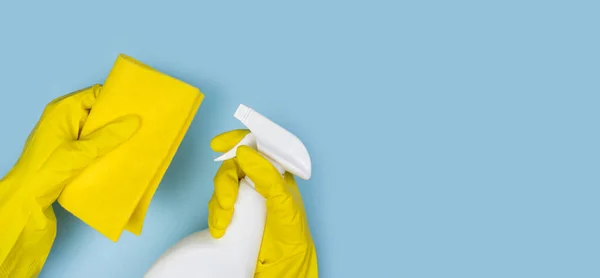 Woman\'s hands in yellow rubber gloves hold a window cleaner spray and rag on a blue background. Banner. Close-up. Top view. Copy space.