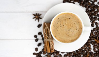 Bir fincan taze kahve, tarçın çubukları ve beyaz ahşap arka planda dağınık kahve çekirdekleri. Kahve günü. Yakın plan. Üst Manzara. Mesaj için yer. Seçici odak.