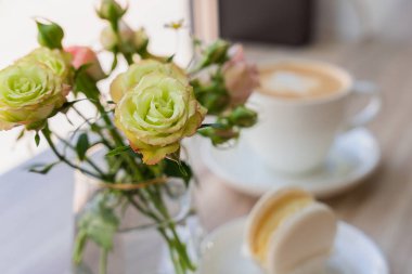 Vazoda bir buket gül ve bulanık bir vazoda bir fincan kapuçino ve bir kafedeki masada tatlı. Yakın plan. Seçici odak.