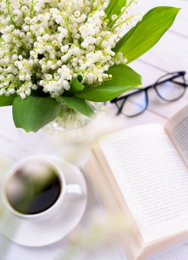 Bir buket taze vadi zambağı, bir fincan sabah kahvesi, masada kitap ve bardaklar. Seçici odak.