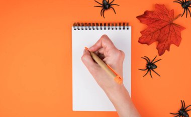 Cadılar Bayramı için yaratıcı kompozisyon. Bir kadının eli, portakal rengi arka planda örümceklerle birlikte deftere bir şeyler yazar. Flatlay. Üst Manzara. Uzayı kopyala Seçici odak.