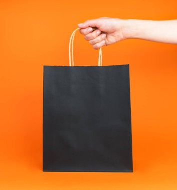 Bir kadının eli turuncu arka planda siyah bir alışveriş çantası tutuyor. Sonbahar indirimi. Cadılar Bayramı alışverişi. Model yap. Boşluğu kopyala.