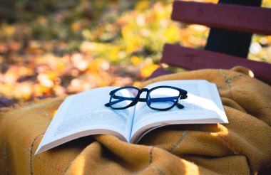 Sonbahar parkındaki bir bankta sıcak bir battaniyeye yerleştirilmiş gözlüklü açık bir kitap. Samimi düşüş atmosferi, okuma, çalışma ve rahatlama konsepti. Seçici odak.