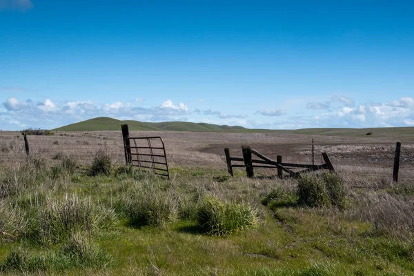 An open gate in a pasture with mostly brown grass indicating the end of the grazing season in Californias Central valley with rolling hills in the background and a mostly cloudless blue sky space for copy