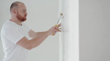 Genç bir adam, duvarı beyaz boyayla silindirle boyuyor. Ev tadilatı konsepti, apartmanda tadilat...
