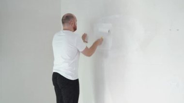 Kafkasyalı bir adam onu yenilemek ve lekeleri kapatmak için duvar boyuyor. Bir ressam elindeki silindiri kullanarak beyaz renk boya kullanarak desenli bir duvarı beyaz renge boyar. Yenileme konsepti. Yüksek kalite