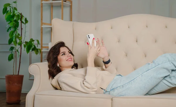 Young beautiful woman sitting on the sofa at home chatting and surfing the net. Female person having fun with smartphone online.