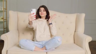 A girl sitting at home on her sofa, having a conversation over a distance call on her smartphone with a big smile on her face, visibly happy to be at home.