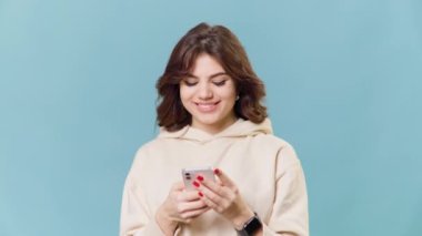A teenage girl in a studio shot, sitting against a bright blue background. She holds a smartphone in her hand and makes a satisfied and accepting gesture while looking at the camera,