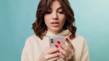 A pretty teenager in a studio shot, laughing and looking at camera joyful as she shops on her smartphone. Her purchase is astonishing and the way she is enjoying it is stunning.