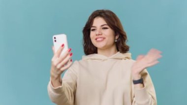 A young adolescent girl is seen in the photo, sitting on a blue background in a studio. She is a blogger and vlogger, and is captured in a moment of happy conversation and greeting,