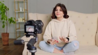 smiling teenager at home, sitting on her sofa and talking to a friend over a video call while gesturing and speaking about her experiences as a student, content creator, and successful YouTuber.