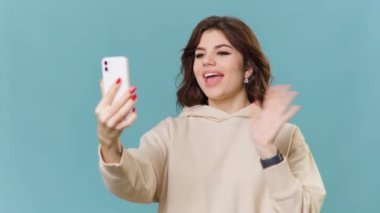 A teenage girl with brunette hair is captured in a studio shot, sitting against a bright blue background. She is a blogger and vlogger, and is seen using her smartphone while smiling and talking