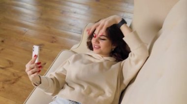 A brunette woman sitting comfortably at home on her sofa, her face alight with happiness as she talks to a friend over a video call on her smartphone.