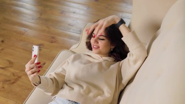 一位黑发女子舒适地坐在沙发上 在用智能手机与一位朋友交谈时 脸上洋溢着幸福的表情 — 图库视频影像