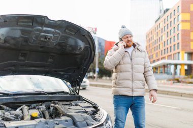 A lone man, looking unhappy and stressed, stands by his damaged vehicle on the side of the road, holding his mobile phone and trying to find a solution to his transportation problem.