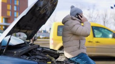 A lone man, looking unhappy and stressed, stands by his damaged vehicle on the side of the road, holding his mobile phone and trying to find a solution to his transportation problem.