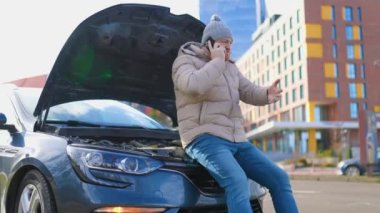 A worried driver is seen holding his mobile phone and standing alone next to his broken down car on the side of the road, possibly calling for emergency assistance or insurance.