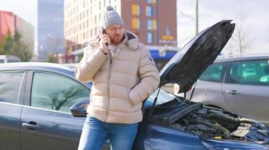 A worried and stressed driver leans against his broken car, holding his mobile phone to his ear as he speaks to someone, likely seeking assistance for his vehicle failure.