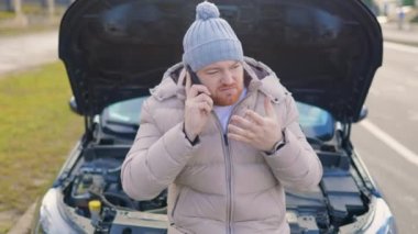 A man is seen on the phone, possibly calling for a tow truck for his broken down car on the side of the road, with a look of despair on his face.