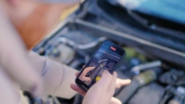 top view of a man with smartphone examines the damage engine, likely trying to find help for his vehicle breakdown. receives remote assistance