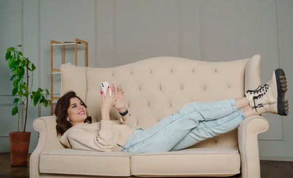 Young beautiful woman sitting on the sofa at home chatting and surfing the net. Female person having fun with smartphone online.