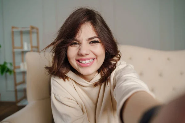 Pretty young asian female with big smile sitting at living room. She having fun taking light cheerful selfie on blurred background. High quality photo
