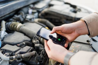 close up a man on the side of the road, using his mobile phone to call for help as he inspects the damage to his vehicles engine, while his car remains in a broken state.