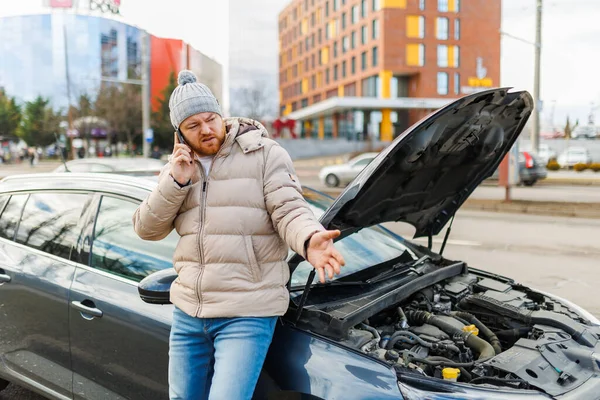 A worried and stressed driver leans against his broken car, holding his mobile phone to his ear as he speaks to someone, likely seeking assistance for his vehicle failure.