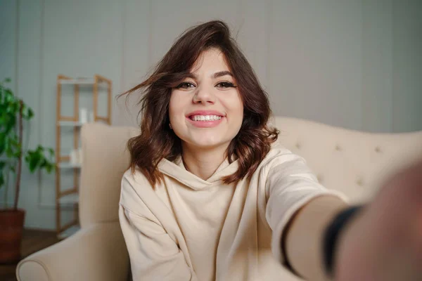 Pretty young asian female with big smile sitting at living room. She having fun taking light cheerful selfie on blurred background. High quality photo