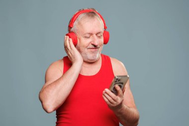 In this studio shot on a blue background, an elderly man enjoys a good laugh as he listens to music through his headphones and uses his smartphone to play an app. joyful experience of seniors