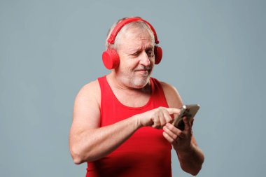 A laughing senior man with a satisfied expression listens to music with headphones. The studio shot features the man on a blue background,