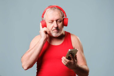 In this studio shot on a blue background, a senior man uses his smartphone with headphones in hand as he listens to music. technology usage for older generations.