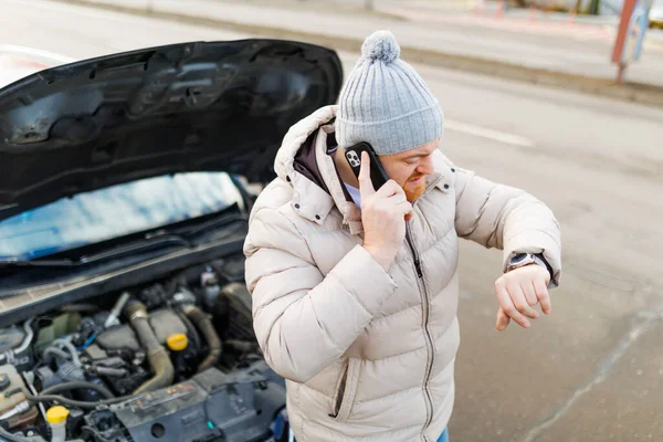 A young man stands alone on the side of the road, looking dejected as he holds his smartphone and talks to someone, likely seeking help with his broken-down car.