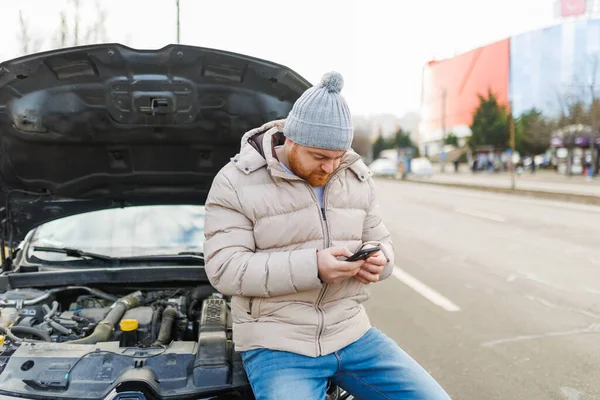 A worried and stressed driver leans against his broken car, holding his mobile phone to his ear as he speaks to someone, likely seeking assistance for his vehicle failure.