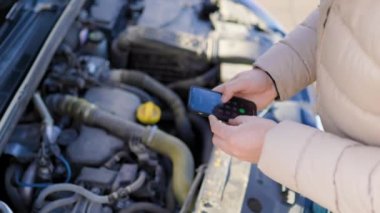 A close-up of a drivers hands holding a smartphone, calling for roadside assistance as he tries to repair his broken down car.