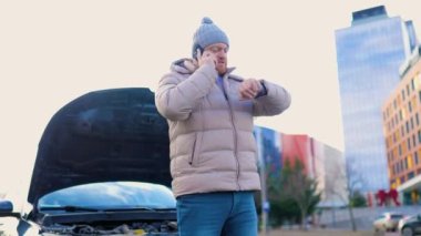 A forlorn man stands beside his broken car, with a troubled expression as he speaks on his phone, seeking assistance for his breakdown.