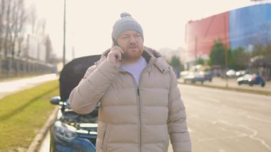Hüsrana uğramış bir sürücü cep telefonunu tutarken ve yolun kenarında bozuk arabasının yanında tek başına dururken görülüyor, muhtemelen acil yardım ya da sigorta yardımı istiyor..