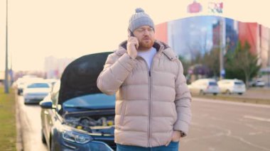 An authentic, real-life image of a man in trouble as he stands by his damaged vehicle on the side of the road, looking angry and upset while talking on the phone, seeking support and assistance