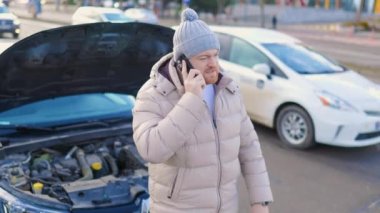 A young man stands alone on the side of the road, looking dejected as he holds his smartphone and talks to someone, likely seeking help with his broken-down car.