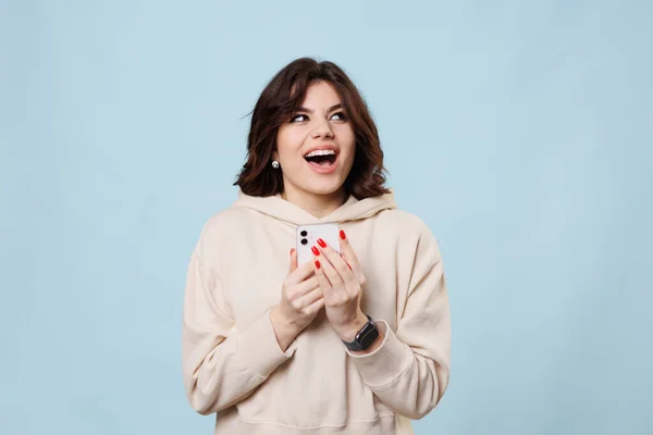 A pretty teenager in a studio shot, laughing and looking at camera joyful as she shops on her smartphone. Her purchase is astonishing and the way she is enjoying it is stunning.