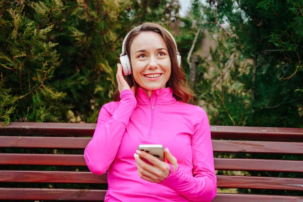 Güzel, genç bir kadın, esmer saçlı, park yerinde tek başına oturuyor, güneşin tadını çıkarıyor ve kulaklıklarıyla müzik dinliyor. Aktif ve tasasız bir yaşam tarzının mükemmel bir vücut bulmuş hali.
