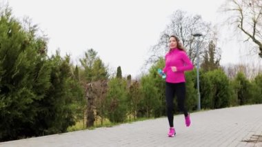 A determined female runner jogging in the park. A sporty hispanic woman in a pink sportswear set jogging on the street, holding a water bottle